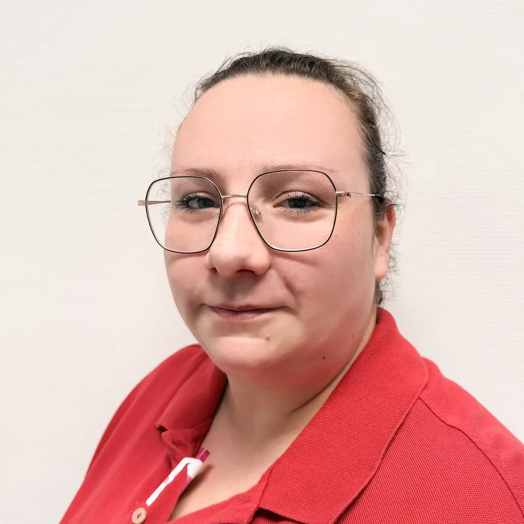 Eine laechelnde Frau mit Brille und rotem Poloshirt vor hellem Hintergrund